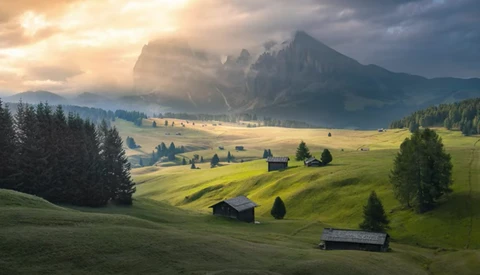 Alpine valley with scattered wooden chalets, rolling green meadows, and dramatic rocky peaks under golden sunrise light.