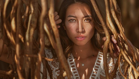 Woman with dark hair framed by dried grass, wearing a white textured top and looking directly at camera.