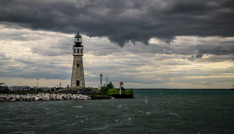 Lighthouse standing on rocky breakwater under dramatic storm clouds with choppy water in foreground.