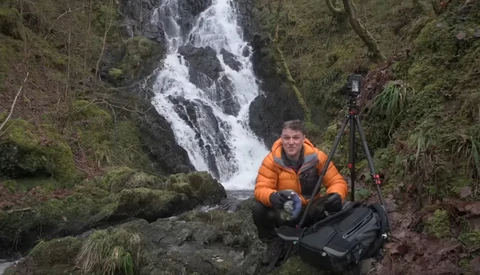 Photographer in orange jacket crouching by waterfall with camera gear in forested landscape.