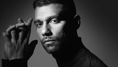 Black and white portrait of a bearded man with hand raised to his face, studio lighting against neutral background.