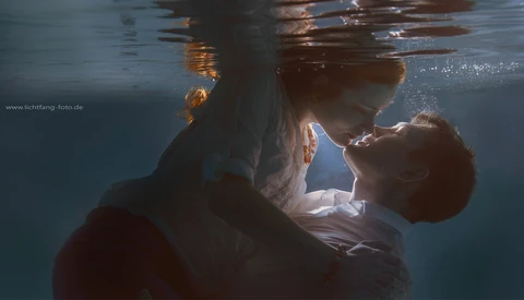 Two people embracing underwater in shallow water with natural sunlight filtering from above.