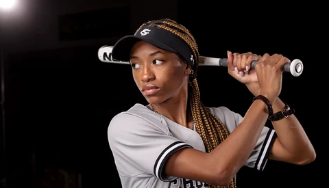 Female athlete in baseball uniform holding bat over shoulder against dark background.