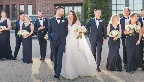 Wedding party lined up outdoors with bride and groom in center, bridesmaids and groomsmen flanking them in formal attire.