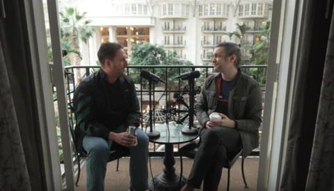 Two men having a conversation on a balcony overlooking a European-style urban streetscape.