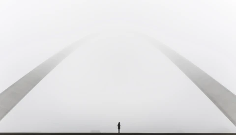 Silhouette of a solitary figure standing beneath a monumental curved architectural structure against an overcast sky.