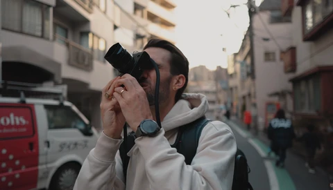 Male photographer holding camera up to his face on an urban street lined with buildings.