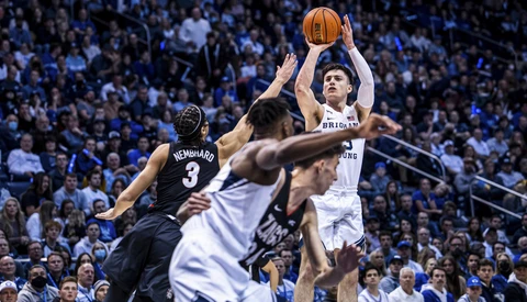 Basketball player in white jersey shooting over defender in black jersey during game with crowd in background.