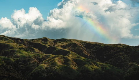Rolling green hills beneath a blue sky with white clouds and a rainbow in the distance.