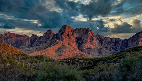 Desert mountain landscape at sunset with layered rock formations and saguaro cacti under dramatic clouds.