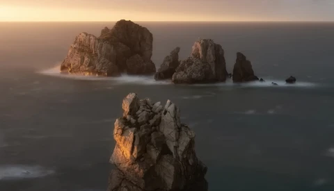 Rocky sea stacks silhouetted against golden sunrise light over misty ocean waters.