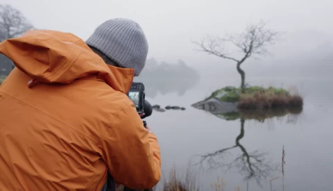 Photographer in orange jacket shooting a minimalist landscape with a solitary tree reflected in still water on a foggy morning.