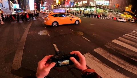 Hands holding a smartphone capturing an orange taxi cab at a busy city intersection at night.