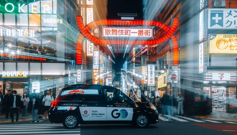 Taxi parked in busy Japanese street intersection at night with large red neon signage overhead.