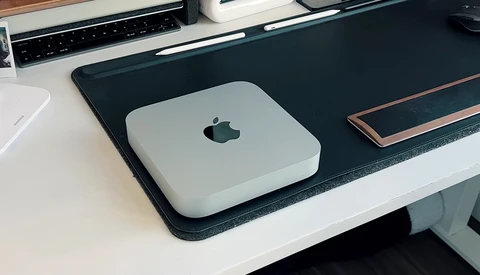 Mac mini computer on a black desk pad next to a keyboard and other workspace items.