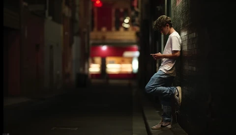 Man leaning against brick wall at night checking mobile phone on dimly lit urban street.