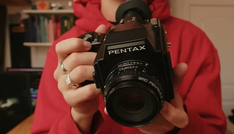 Person in red shirt holding a black Pentax film camera with SMC Pentax lens toward the camera.