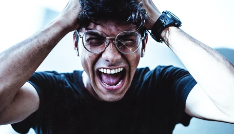 Man wearing glasses with hands on head, smiling widely with an energetic expression against a blue background.