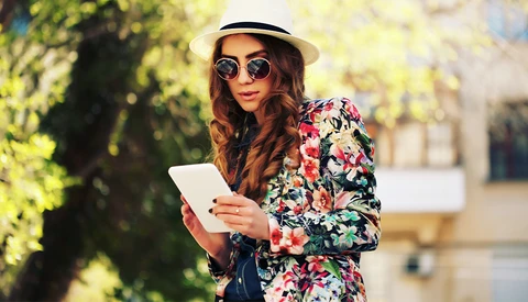 Woman in floral blazer wearing sunglasses and straw hat, holding a tablet outdoors.