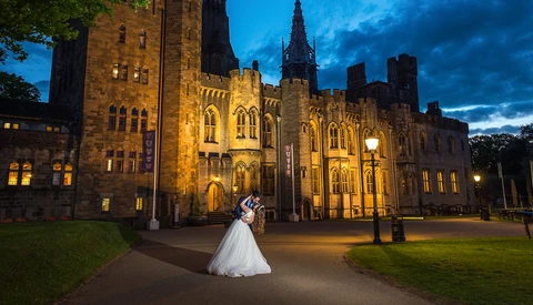Bride and groom posing in front of an illuminated Gothic stone castle at dusk.