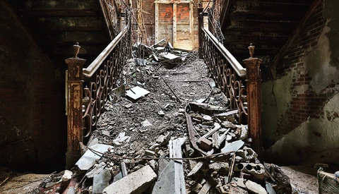 Abandoned stairwell filled with debris and rubble, with ornate metal railings and an open doorway at the far end.