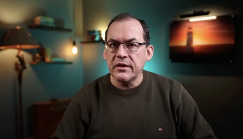 Man wearing glasses and dark sweater speaking in a studio setting with teal walls and ambient lighting.