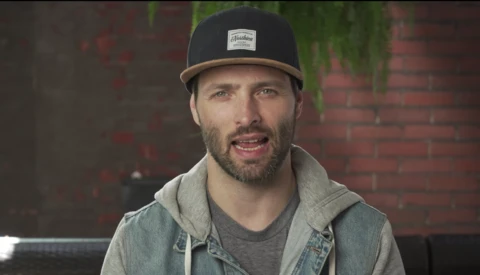 Man wearing a black and tan snapback cap and denim vest over gray hoodie, smiling at camera against brick wall.