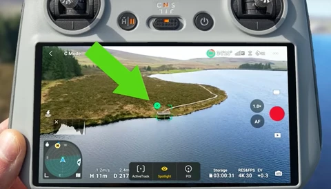 Screenshot of drone controller display showing aerial video feed of a curved shoreline with green arrow pointing to waypoint marker.