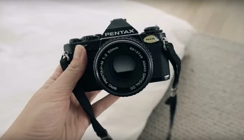 Pentax film SLR camera held in hand with 50mm lens attached and shoulder strap visible.