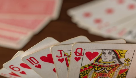 Playing cards fanned out showing hearts suit and a queen card, with additional cards blurred in the background on a dark wooden surface.