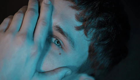 Close-up portrait of a person's face partially obscured by their hand, lit with cool blue and teal tones against dark background.