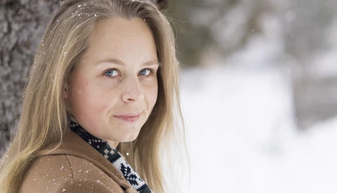Young woman with long blonde hair wearing a dark patterned scarf, photographed outdoors in falling snow.