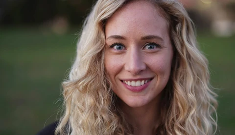 Woman with wavy blonde hair smiling at camera outdoors with blurred green background.