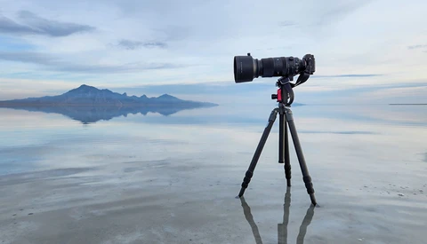 Camera with telephoto lens mounted on tripod at shallow water's edge, photographing distant volcanic island under overcast sky.