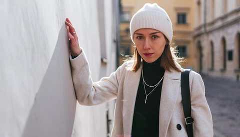 Woman in cream blazer and white beanie poses against white wall in urban courtyard.