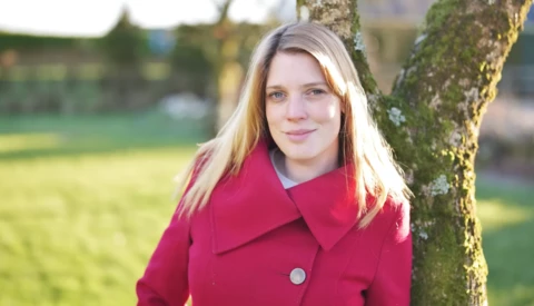 Young woman with long blonde hair wearing a bright magenta coat, posing against a moss-covered tree trunk in an outdoor garden setting.