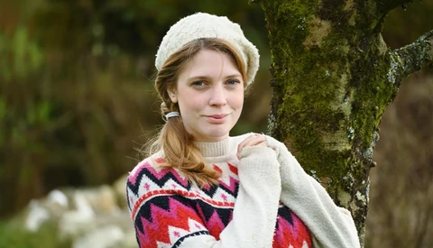 Young woman in cream knit beanie and red patterned sweater leaning against moss-covered tree trunk.