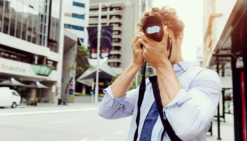 Man holding vintage camera up to his face on an urban street with tall buildings behind him.
