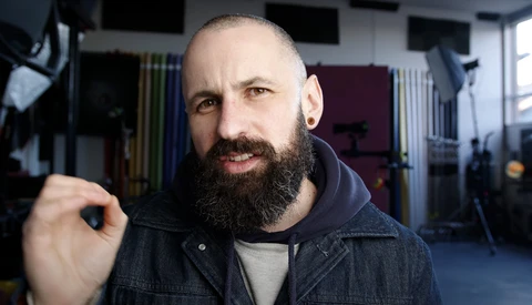 Bearded man with shaved head in denim jacket gestures while speaking in a studio setting.