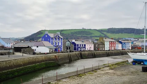 Colorful waterfront buildings reflected in calm harbor water, with sailboat and hillside backdrop.