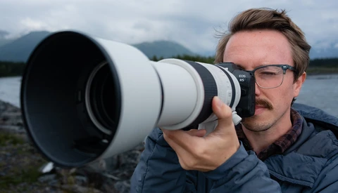 Photographer holding a white telephoto lens toward camera while wearing glasses and dark jacket by a lake.
