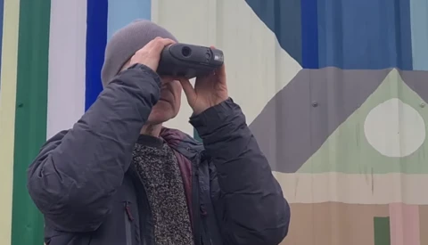 Photographer holding a camera to their face while standing in front of a colorful geometric mural.