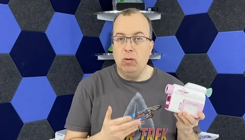Man holding a pink and white device while gesturing during an explanation, wearing glasses and a graphic t-shirt.