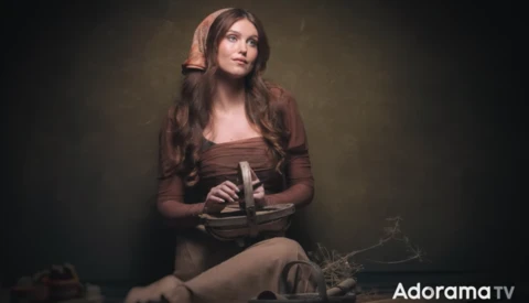 Woman in brown clothing holding a book, posed against a dark studio background with warm lighting.