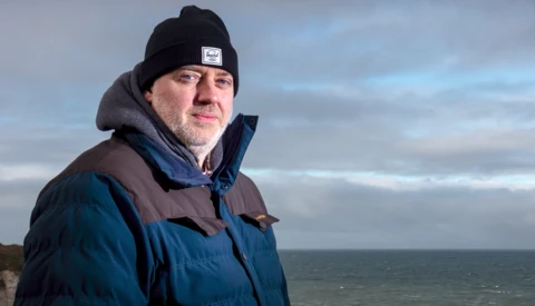 Man wearing blue jacket and black beanie standing outdoors with ocean horizon behind him.