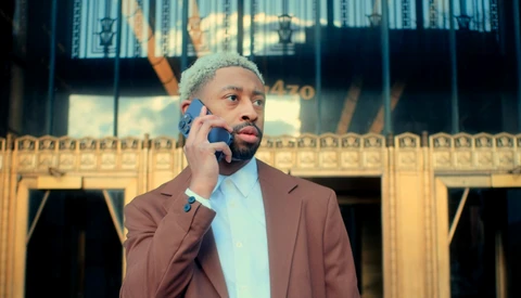 Man in brown blazer and light blue shirt talking on a blue phone with ornate golden building facade in background.