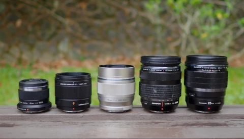 Five camera lenses arranged in increasing size order on a wooden surface against a blurred natural background.