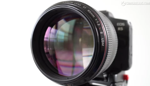 Close-up of a fast telephoto lens mounted on a camera body, showing internal glass elements with purple and magenta lens coatings.