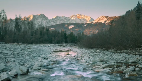 Mountain river flowing through a forested valley at sunrise, with snow-covered peaks glowing in golden light.