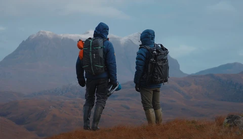 Two hikers with backpacks stand on a ridge overlooking mountainous terrain under partly cloudy skies.